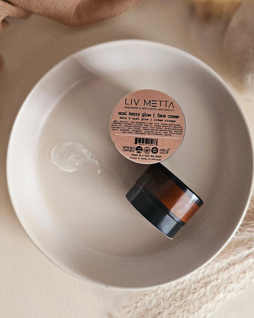 A jar of Acai Berry Glow Face Cream by LIV METTA is placed on a white plate with a lid beside it, on a wooden surface.