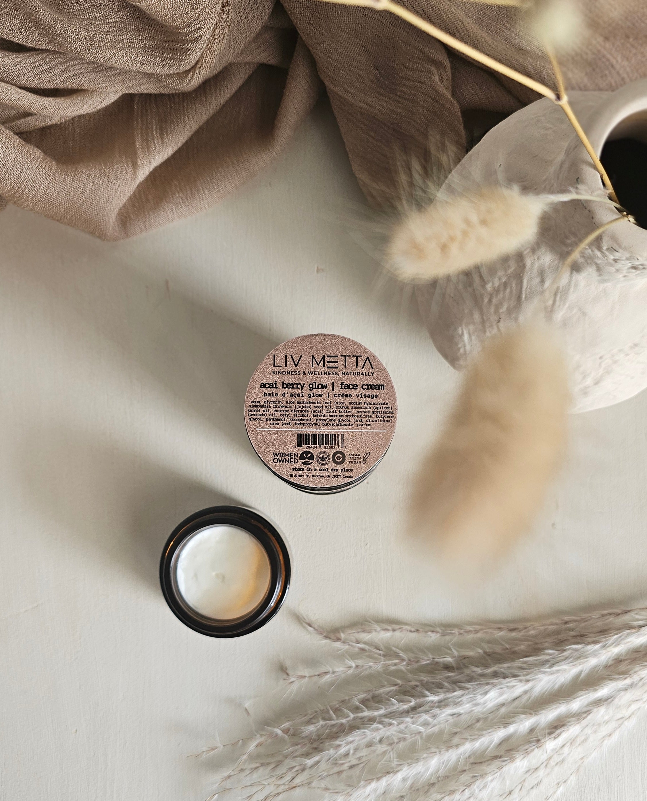 A small jar of Acai Berry Glow Face Cream by LIV METTA is placed on a light-colored surface, partially covered by a beige cloth. The jar is open, showing the cream inside, and there are decorative dry plants next to it.