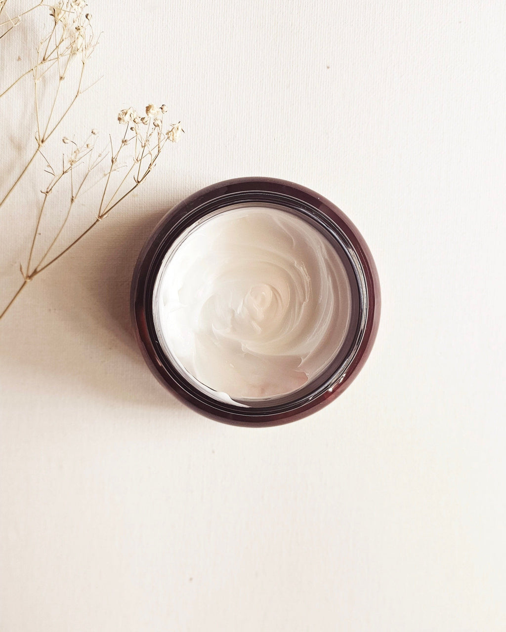 A jar of Aromatic Body Lotion with a amber color lid, placed on a light background, with a sprig of dried flowers to the left.