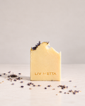 A single bar of pale yellow soap standing upright on a light surface, with a bite taken out of the top and dried lavender sprigs resting on the edge. The soap is embossed with the brand name 'LIV METTA', and scattered dried lavender is visible around it.