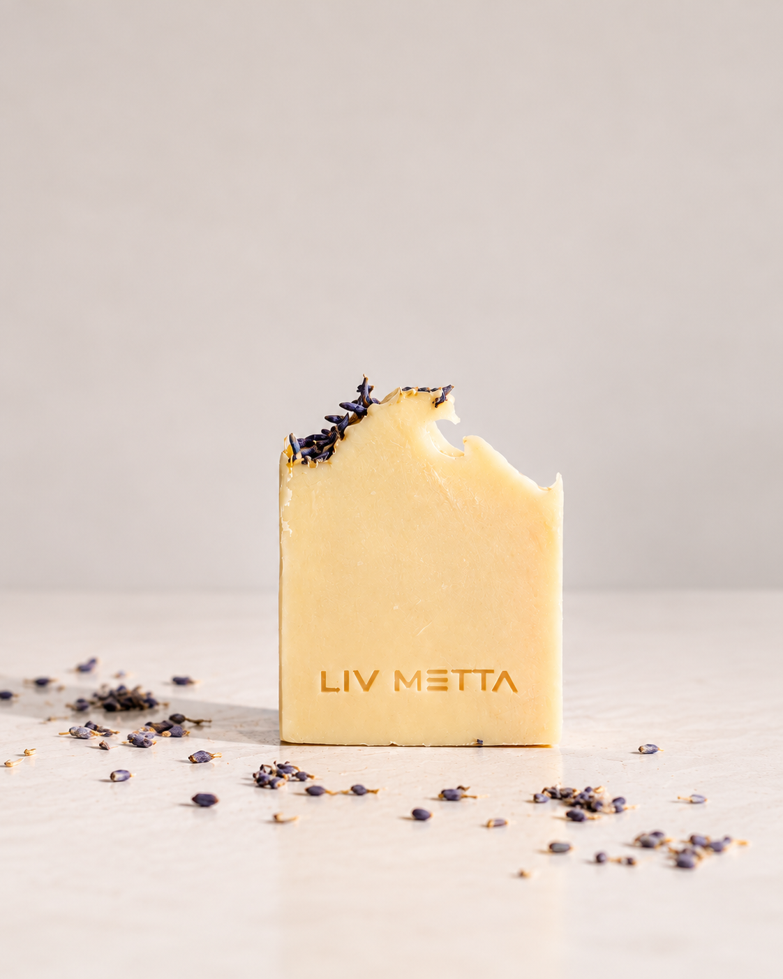 A single bar of pale yellow soap standing upright on a light surface, with a bite taken out of the top and dried lavender sprigs resting on the edge. The soap is embossed with the brand name 'LIV METTA', and scattered dried lavender is visible around it.