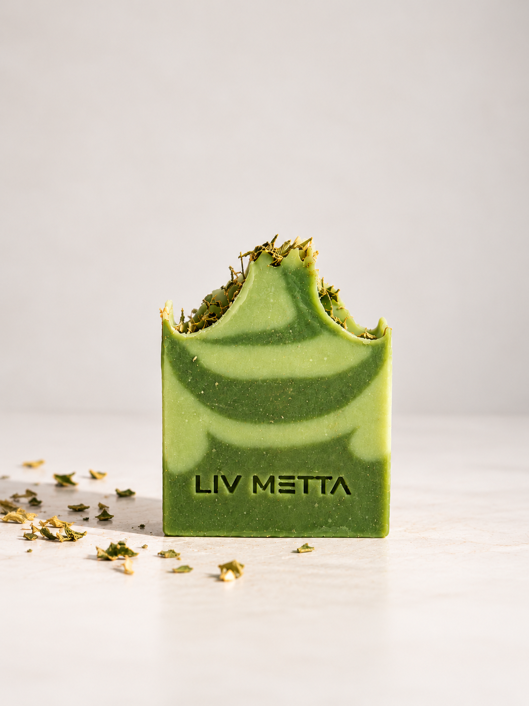 A rectangular bar of green soap with a bite taken out of the top, embossed with the brand name 'LIV METTA', and topped with dried green herbs, set against a clean light gray background with scattered herb pieces on the surface.