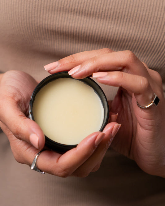 A person holding a small open jar of moisturizing hand and body balm in their palms.