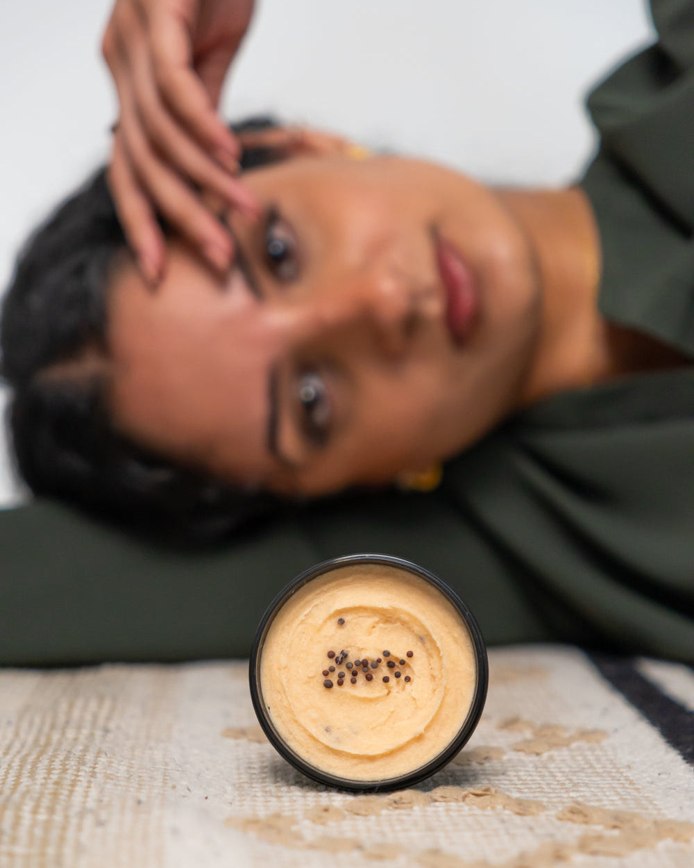 A jar of sugar body scrub with a creamy texture, with a model lying down in the background.