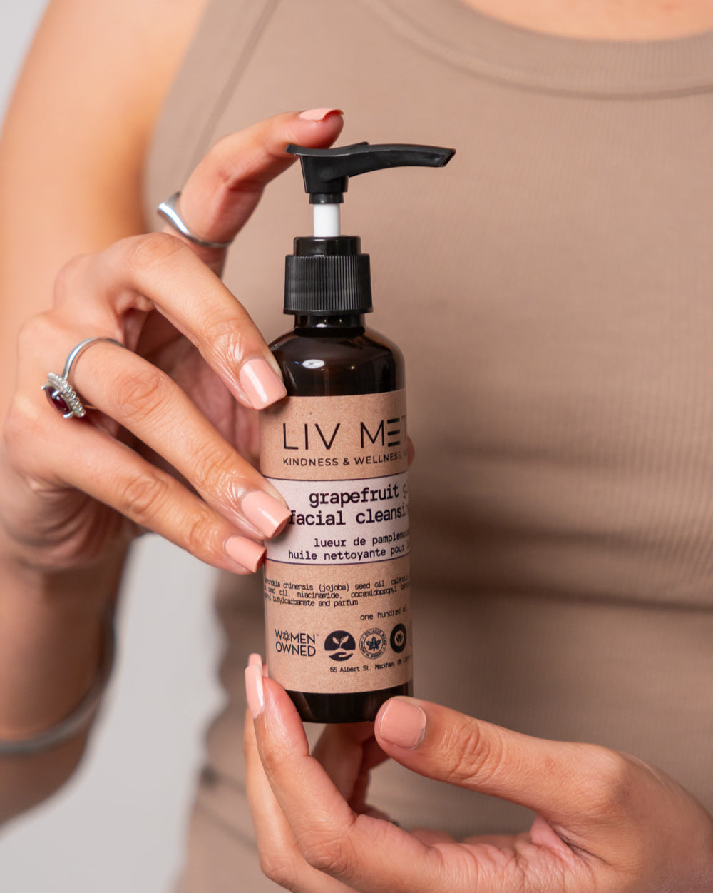 A person holding a bottle of Grapefruit Facial Cleansing Oil with a pump dispenser. The label includes the brand name 'LIV META' and the product name 'grapefruit glow facial cleansing oil'.