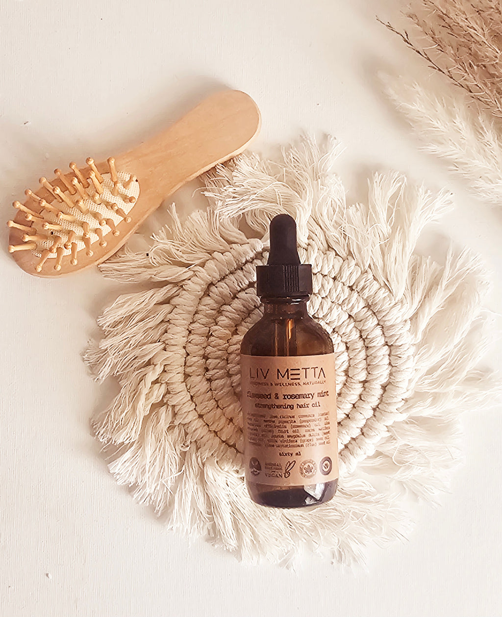 A bottle of Flaxseed & Rosemary Mint Hair Oil with a dropper cap, placed next to a wooden hairbrush, presented on a white textured surface.