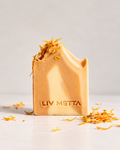 A rectangular bar of LIV METTA soap in a warm golden-beige color, decorated with dried orange peel and standing on a light surface with a few pieces scattered around. The brand name 'LIV METTA' is embossed on the front of the soap against a clean, light gray background.
