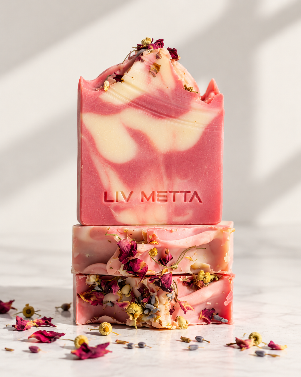 Two stacked bars of LIV METTA handmade soap with a pink and cream marbled pattern, decorated with dried rose petals and botanicals, set on a light marble surface with soft shadows.