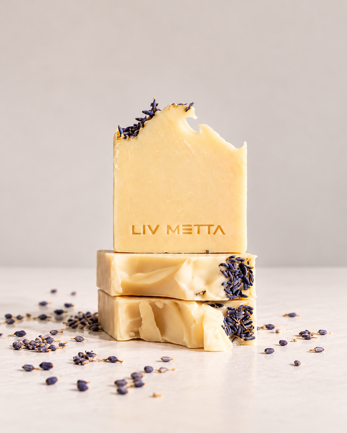 Three stacked bars of handmade soap with lavender buds embedded in them, featuring the brand name 'LIV METTA' on the front bar, set against a neutral light gray background with scattered dried lavender seeds.