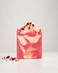 A rectangular bar of handmade soap with pink and white marbled swirls, topped with dried flower petals, standing upright on a light gray surface against a light gray background. The brand name 'LIV METTA' is embossed on the front.