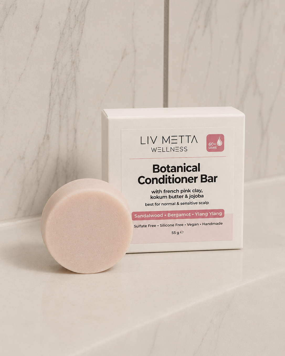 A round pale pink botanical conditioner bar sits in front of its white packaging box on a light marble surface and matching marble background. The box reads 'LIV METTA WELLNESS' and 'Botanical Conditioner Bar'.