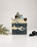 A handmade rectangular bar of soap with irregular rustic broken tops, featuring a marbled swirl of dark gray, cream, and light blue. The brand name 'LIV MEITTA' is embossed on the front.