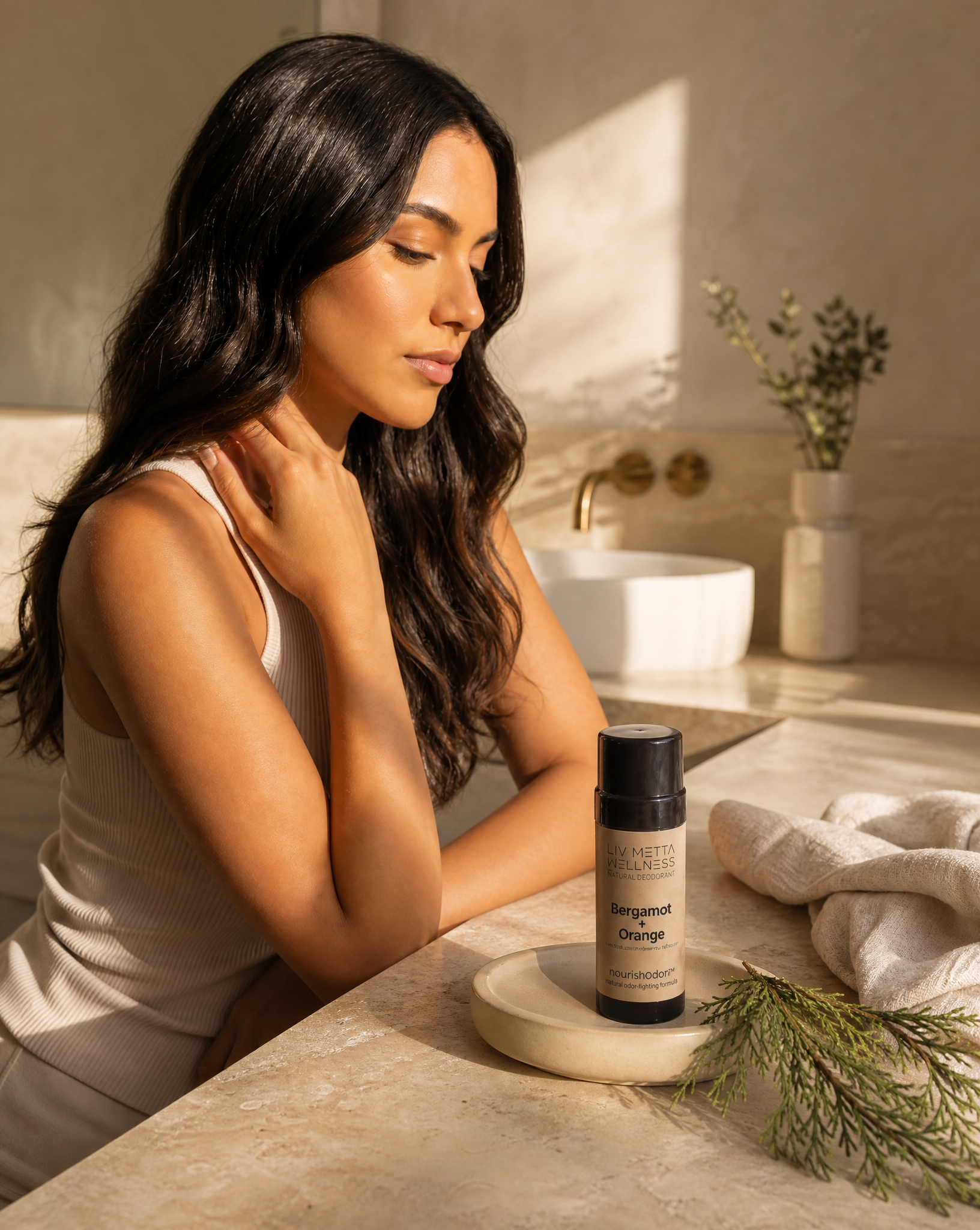 A woman with long dark hair sits at a bathroom counter, looking down at a small bottle of Perugano & Orange hair product placed on a small dish. The scene is warmly lit with sunlight, a towel, and a vase of dried stems in the background.