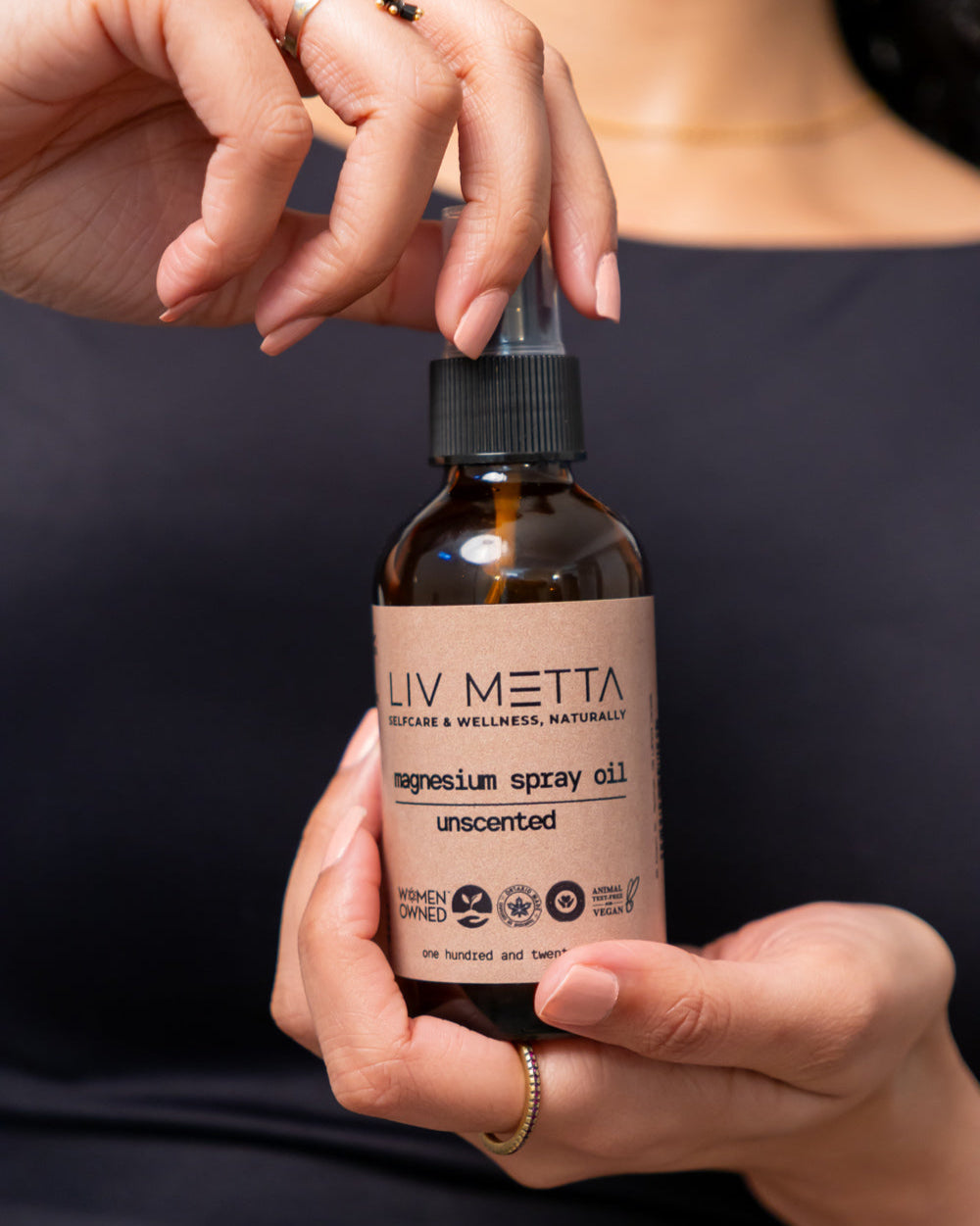 A person holding a bottle of Topical Magnesium Oil Spray with a label that reads 'LIV METTA magnesium spray oil unscented'.