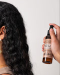 A person with curly hair is shown from the back, with a spray bottle of Rosemary Shea Hair Mist being used on their hair.