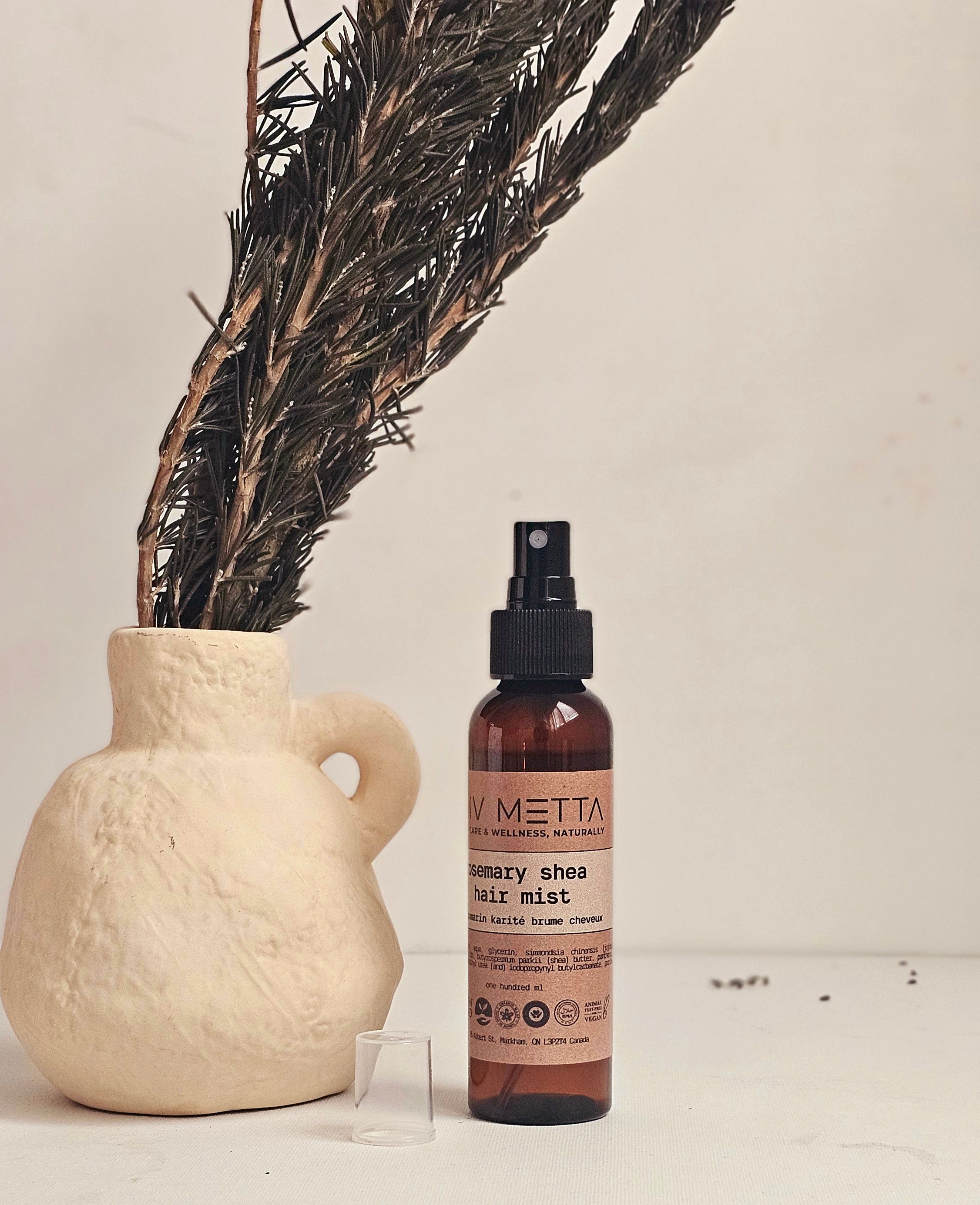 A spray bottle labeled 'Vitessa Rosemary Shea Hair Mist' standing next to a vase with dried rosemary branches, on a white surface.