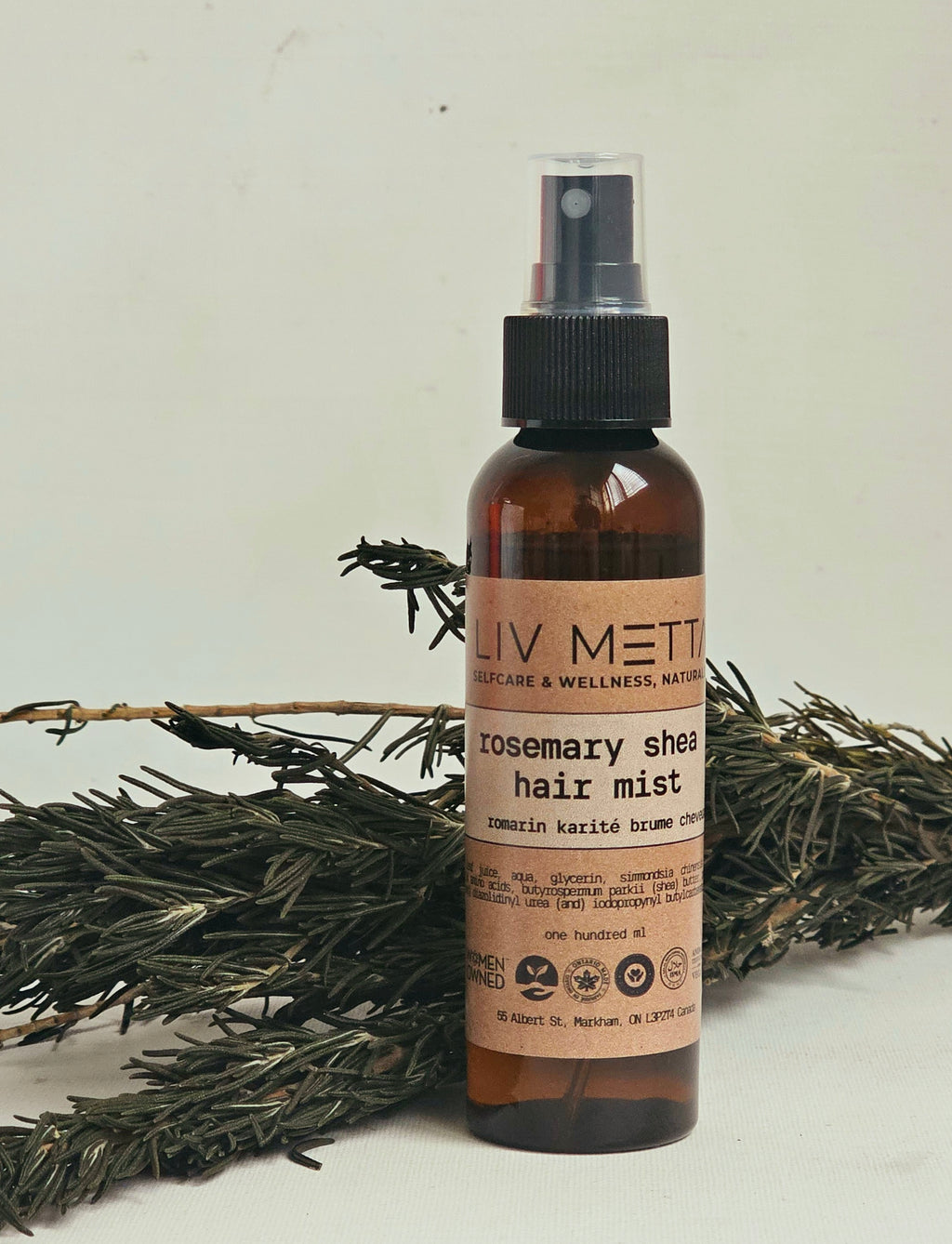 A brown spray bottle labeled 'Rosemary Shea Hair Mist' from the brand LIV MET, positioned next to a sprig of rosemary.