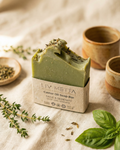 Green soap bar with dried herbs on a textured surface, surrounded by small ceramic containers.