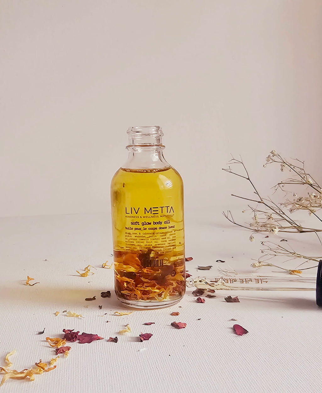A clear glass bottle labeled 'LIV META' filled with a yellow liquid, possibly the Soft Glow Rose Oil, surrounded by dried flowers on a light background.
