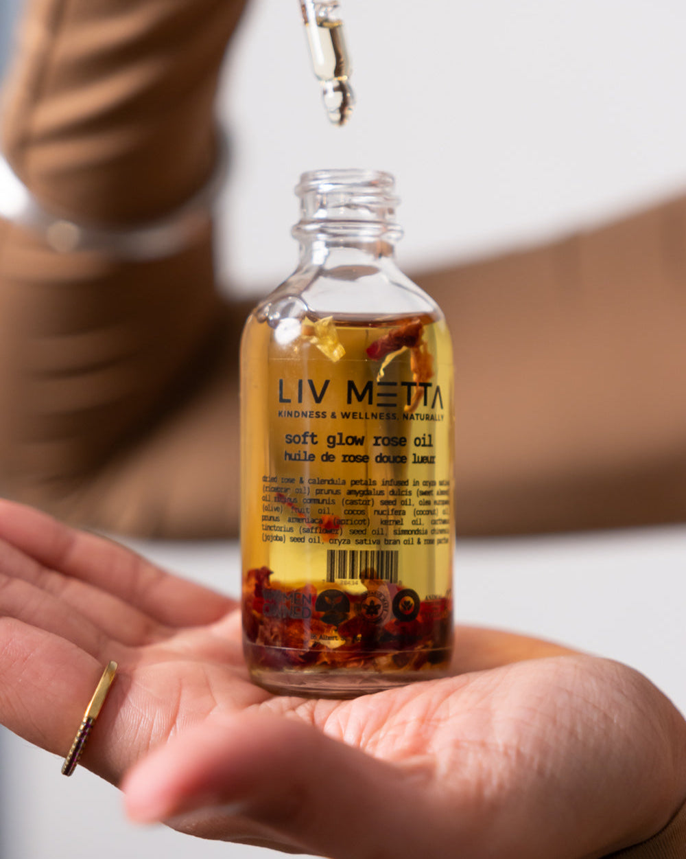 A clear glass bottle with a dropper filled with rose glow oil, held in someone's hand, with visible rose petals inside the bottle.