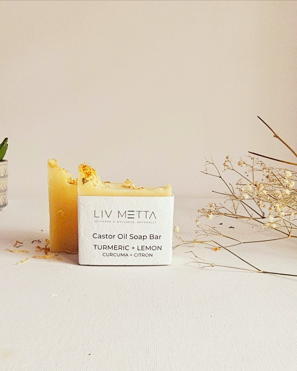 A solid white bar of soap labeled 'LIV METTA Castor Oil Soap Bar' with yellow turmeric and lemon hues on the surface, placed on a white surface with some plant parts in the background.