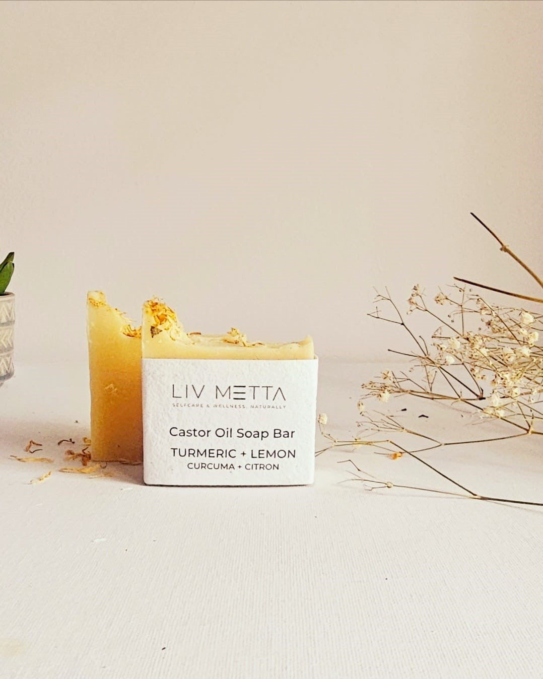 A solid white bar of soap labeled 'LIV METTA Castor Oil Soap Bar' with yellow turmeric and lemon hues on the surface, placed on a white surface with some plant parts in the background.