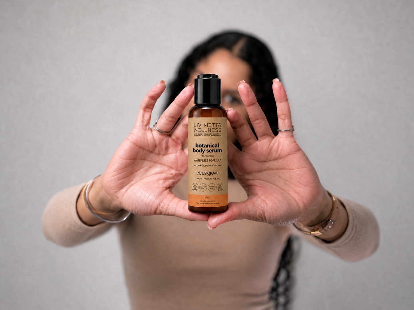 A woman holds a bottle of Lav Mitten Kill Neez Botanical Body Serum in front of her face, showing the amber bottle and black cap against a neutral gray background.